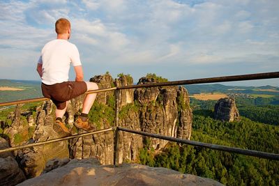 Ginger man sit on handrail at peak of rock and watch sunny day. hiker with shirt, pants and boots.