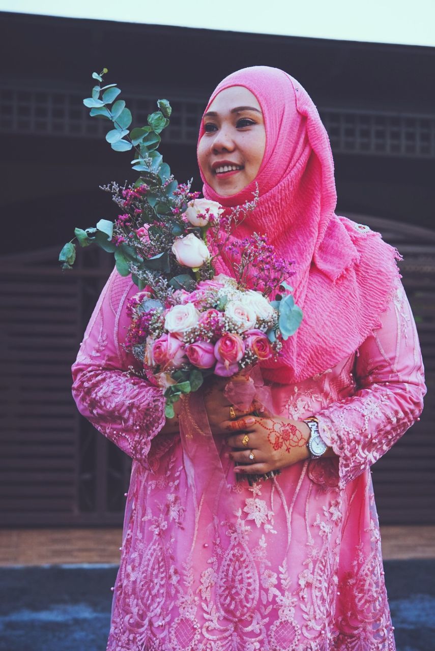 smiling, adult, one person, women, traditional clothing, pink, happiness, clothing, flower, flowering plant, standing, emotion, plant, bride, flower arrangement, nature, celebration, young adult, portrait, three quarter length, lifestyles, front view, bouquet, holding, female, purple, dress, tradition, looking at camera, outdoors, cheerful, day, event, waist up, architecture, person, costume, positive emotion, smile, teeth, wedding dress