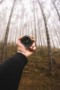 Close-up of hand holding navigational compass against bare trees in forest during foggy weather