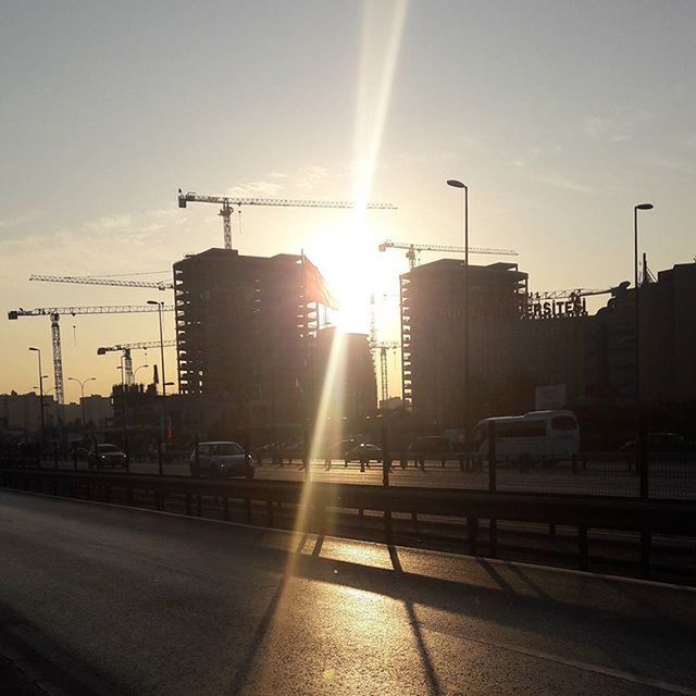 sun, building exterior, built structure, transportation, architecture, sunlight, sunbeam, sky, sunset, mode of transport, lens flare, city, road, street light, car, street, outdoors, silhouette, land vehicle, bright