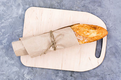 Fresh baguette in wrapping paper on cutting board on table. top view