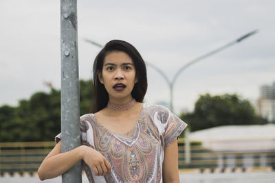 Portrait of surprised young woman holding pole while standing on sidewalk in city