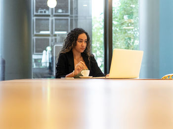 Hispanic young female manager with curly hair stirring coffee in cup and browsing data on netbook while sitting at table and working on remote project in cafeteria