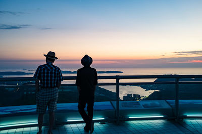 Rear view of men standing on railing against sky during sunset