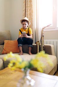 Child in hat text messaging on cellphone while sitting on couch with saxophone in living room