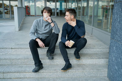 Front view of two young men talking sitting on a city staircase and lo