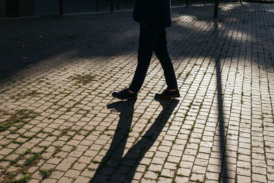 Crop anonymous male in outerwear strolling on paved walkway with shadow on street with bright sunlight in city on sunny day