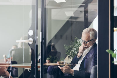 Mature male manager using smart phone seen through glass wall at creative office