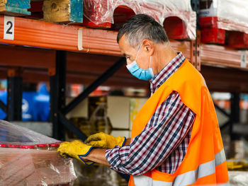 Side view adult professional storehouse specialist in uniform and protective face mask wrapping big carton box while standing near stacks