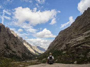 Rear view of man with backpack sitting on rock against sky