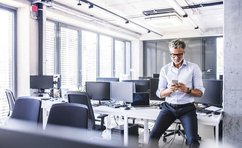 Smiling mature businessman sitting on desk in office using smartphone