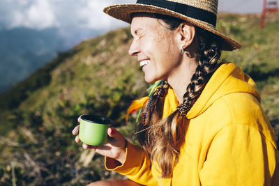 Cheerful mid adult woman wearing hat sitting on mountain during sunny day