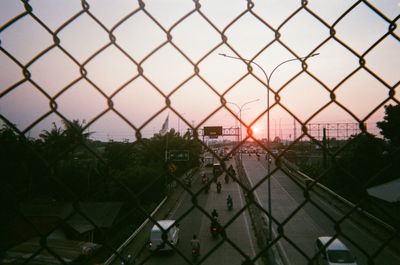 Fence by trees and buildings against sky during sunset