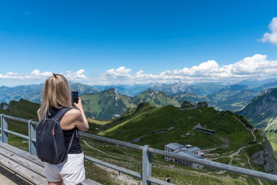 Mature adult blonde woman with backpack taking a photo of the swiss alps from a lookout point.