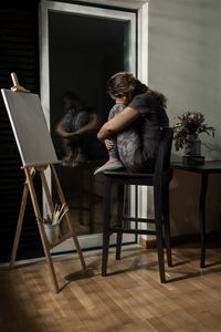 Full length of depressed woman hugging knees while looking through window at home