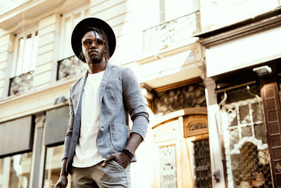 Low angle portrait of young man wearing sunglasses standing against building in city