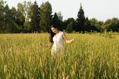 A young woman with long dark hair stands in a field with and looks at the ears of wheat