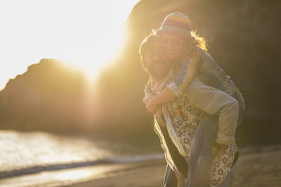 Man piggybacking woman while standing at beach against mountain during sunset