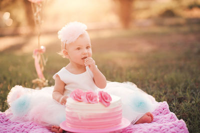 Baby girl 1 year old eating creamy birthday cake sitting on green grass with pink balloons outdoor