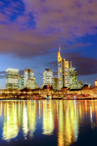 Illuminated buildings by river against cloudy sky