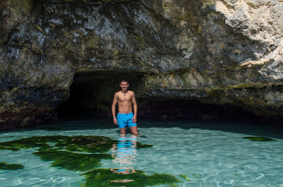 Portrait of shirtless man standing in sea against rock