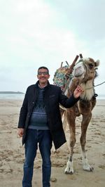 Full length portrait of man standing with camel at beach against sky