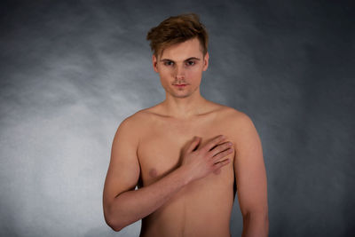 Portrait of shirtless young man with hand on chest standing against gray background