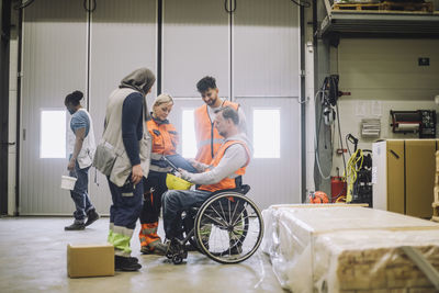 Carpenter sitting on wheelchair discussing over digital tablet with male and female colleagues in warehouse