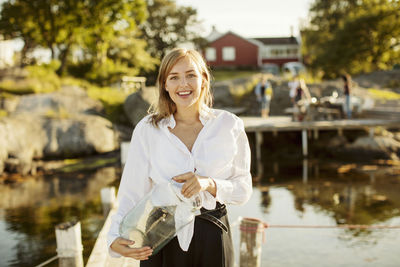 Portrait of smiling young woman holding jar standing on jetty at harbor