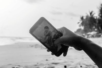 Cropped image of boy clicking selfie with friend photographing while sitting on by sea at beach