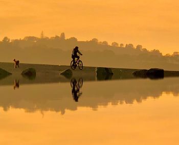 Silhouette man riding bicycle on riverbank against sky during sunset