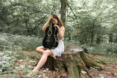 Young woman holding acoustic guitar while sitting on tree stump in forest