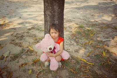Cute girl holding teddy bear while crouching against tree