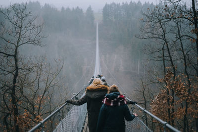 Rear view of people walking on footbridge amidst trees during foggy weather