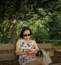 Woman using phone while sitting on bench against trees at park