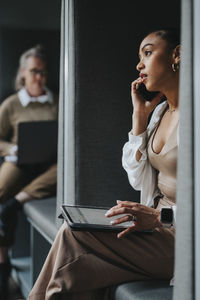 Businesswoman talking on smart phone and using digital tablet while sitting in booth at office