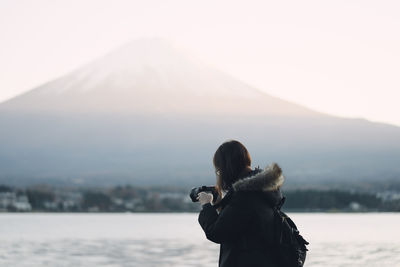 Side view of mid adult woman photographing while standing by lake against mountain