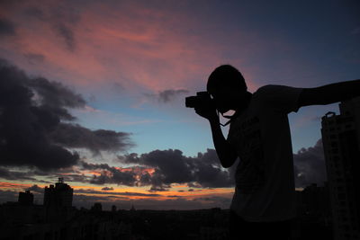 Silhouette man photographing cityscape against sky during sunset