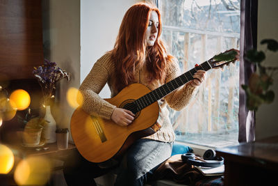 Unaltered candid portrait of young red haired woman in sweater playing acoustic guitar sitting by