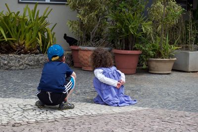 Siblings crouching on footpath against potted plants