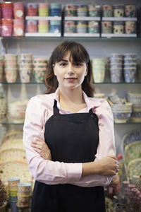 Portrait of confident female owner standing with arms crossed against multi colored crockery at store