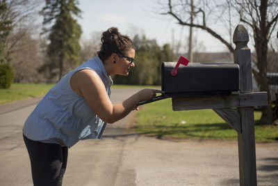 Side view of young woman standing by mailbox on street against sky