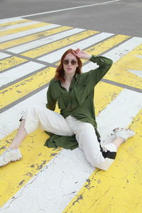 Portrait of ginger woman wearing green shirt and sunglasses with yellow felt hat sitting on road 