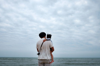 Rear view of father carrying son at beach against cloudy sky