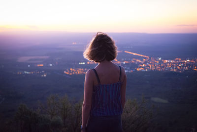 Rear view of woman looking at illuminated cityscape against sky during sunset