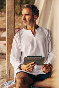 Mature man looking away holding tablet pc while sitting in cabana on vacation at beach