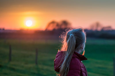 Rear view of girl wearing hooded jacket standing on grassy field during sunset