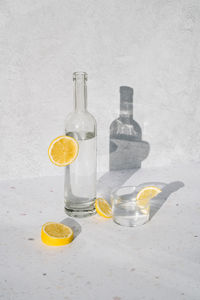 Sunlit glass bottle with water surrounded by lemon wedges on white terrazzo