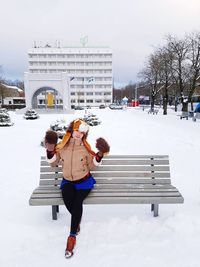 Full length portrait of woman sitting on bench during winter in city