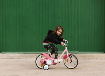 Girl riding bicycle on footpath against green wall